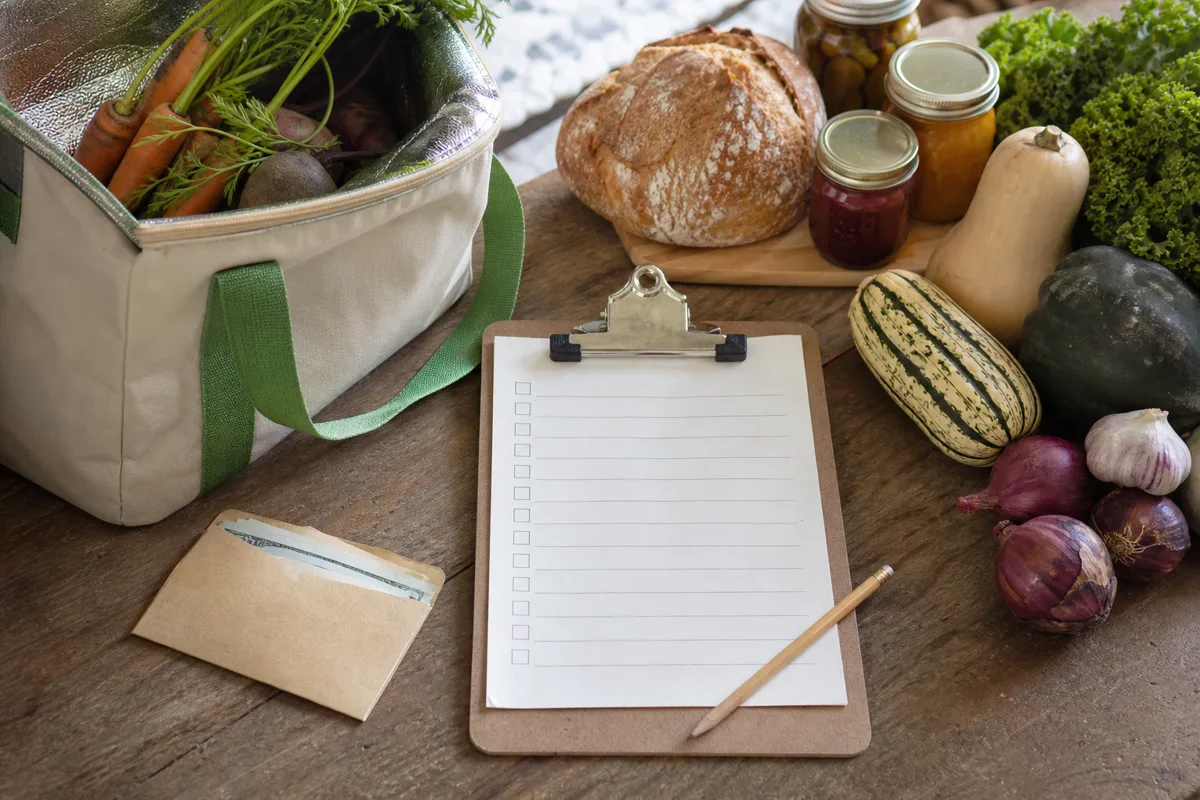 Reusable handwritten market checklist beside vegetables, bread, jars, and an insulated shopping bag
